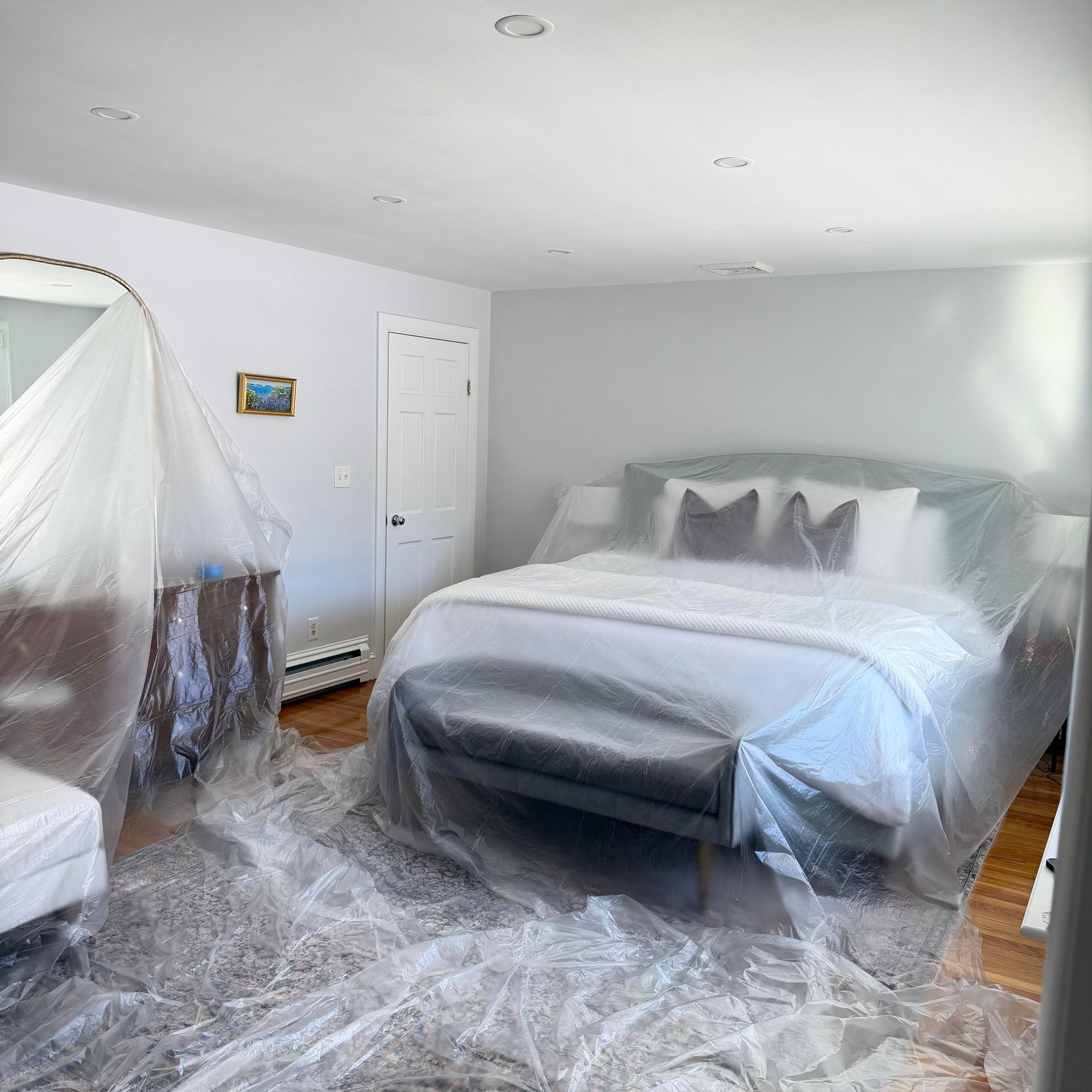 Bedroom with a bed under plastic sheeting, a chair, and a door in a freshly painted room