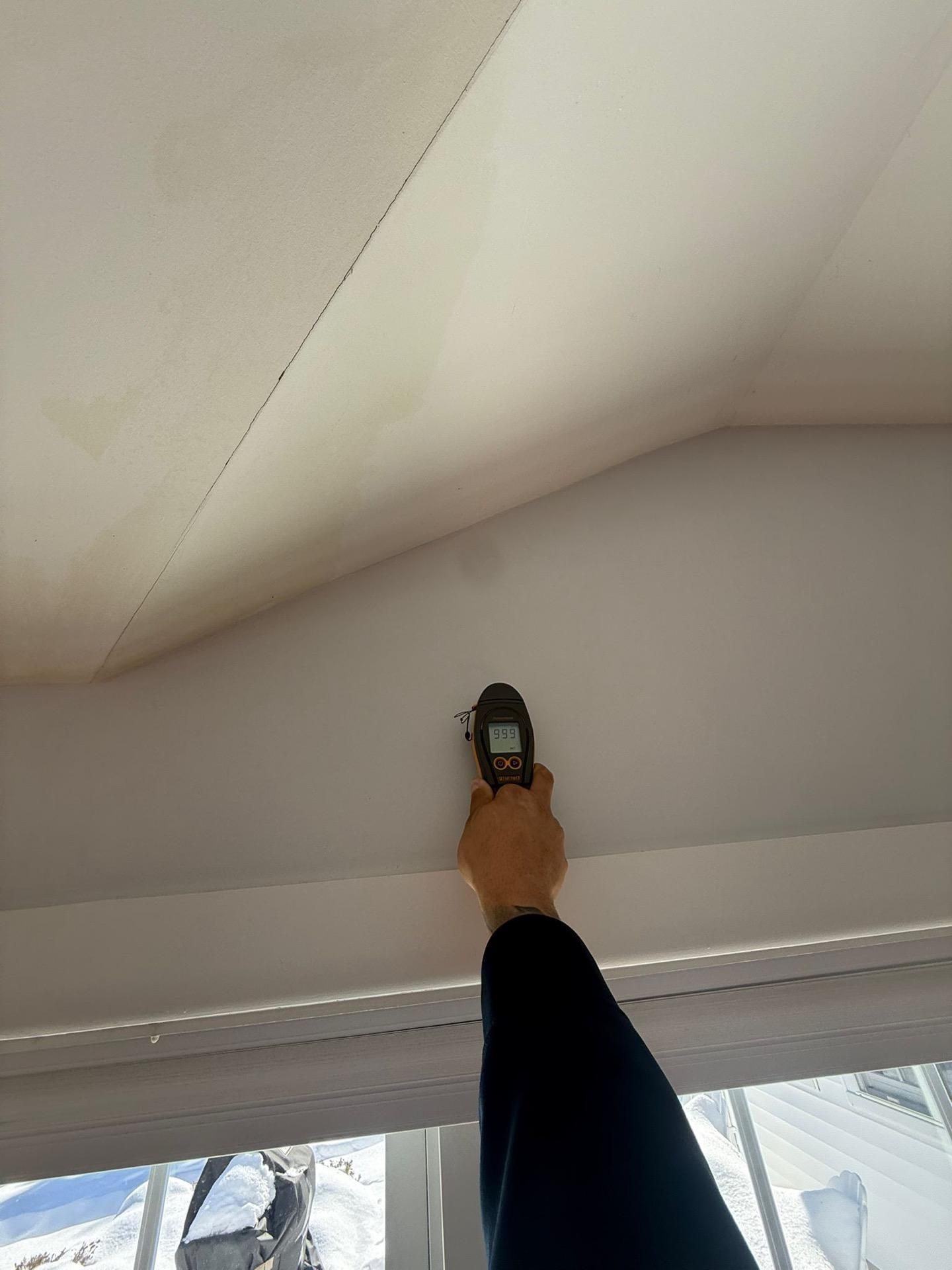 Hand holding a tape measure up to a white ceiling near a window.