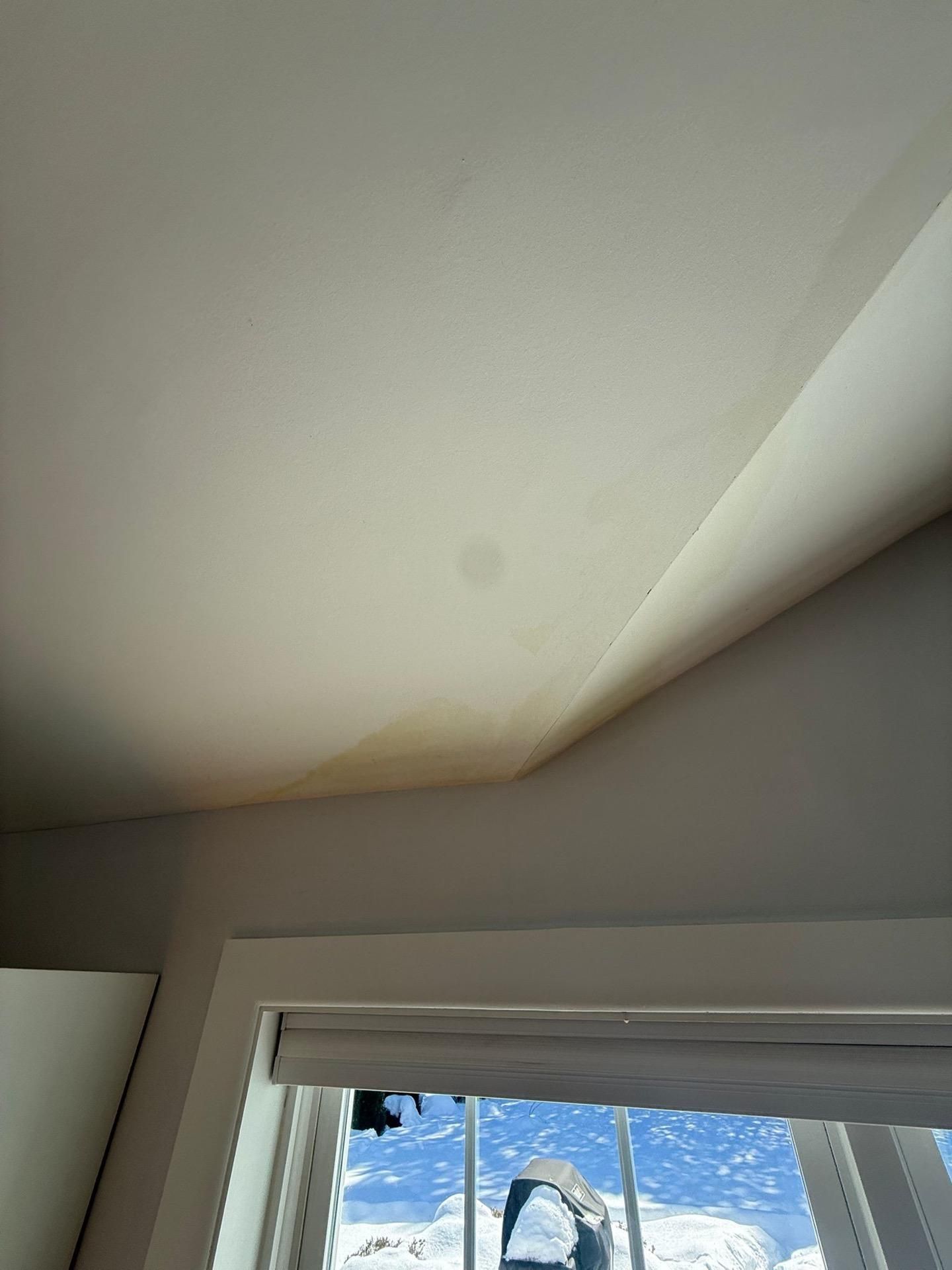 Water-stained ceiling near a window with peeling paint and a blue sky visible outside