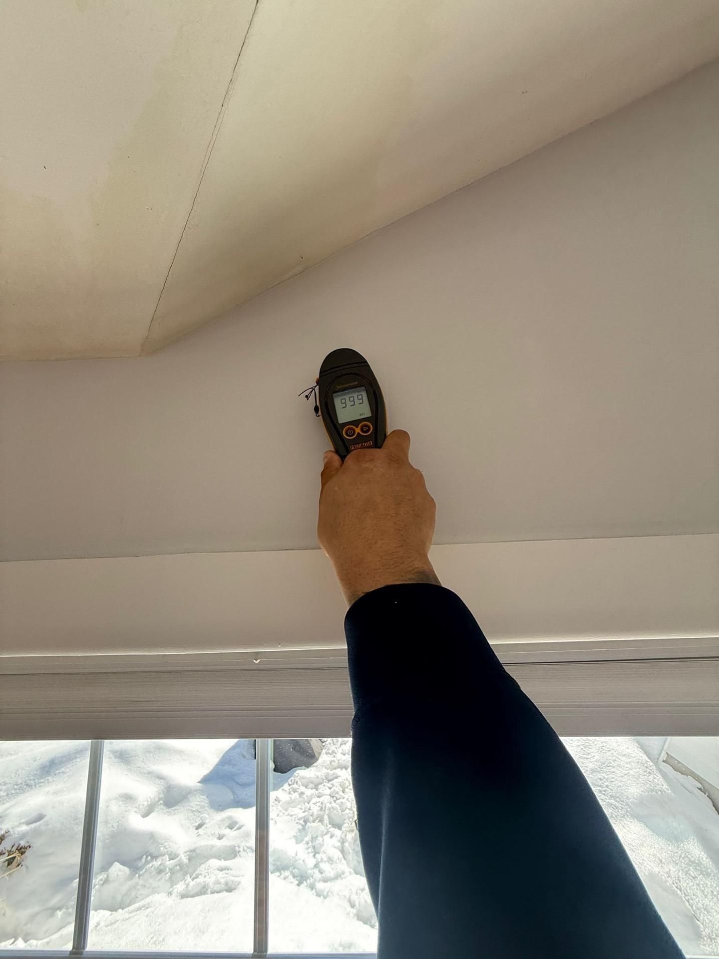 Hand holding a device up to a ceiling seam, measuring moisture near a window.