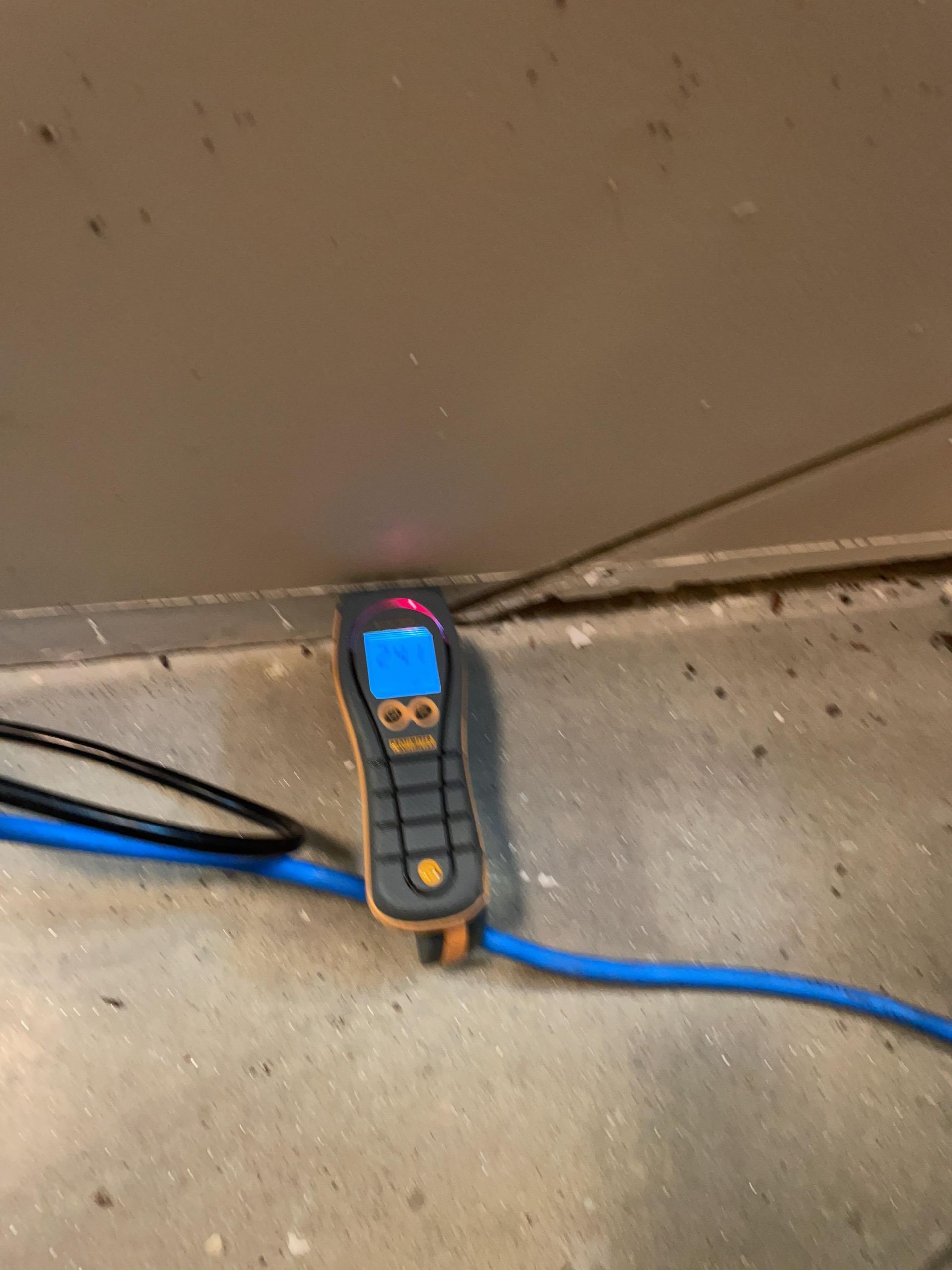 A handheld electronic tester with a blue display rests on a concrete floor beside blue cables.
