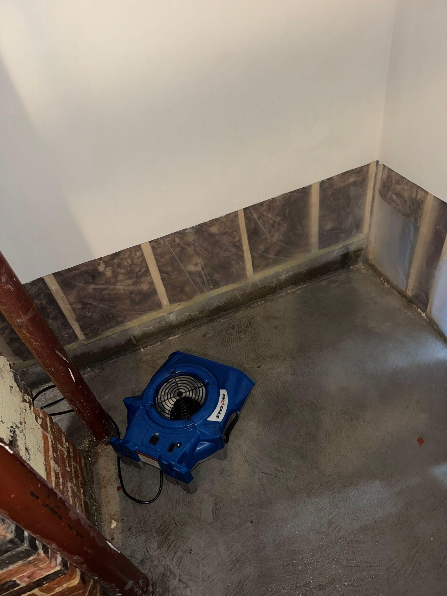 Blue robotic floor cleaner in a corner on a concrete floor near a brick wall.