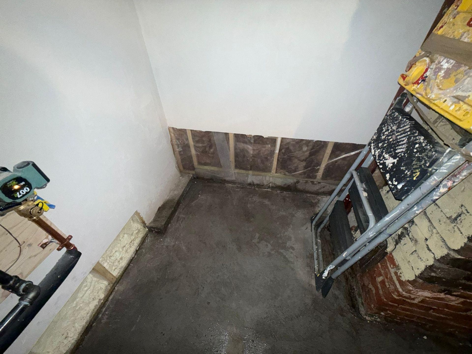 Empty small room corner with white walls, gray floor, and a ladder leaning against a brick wall.