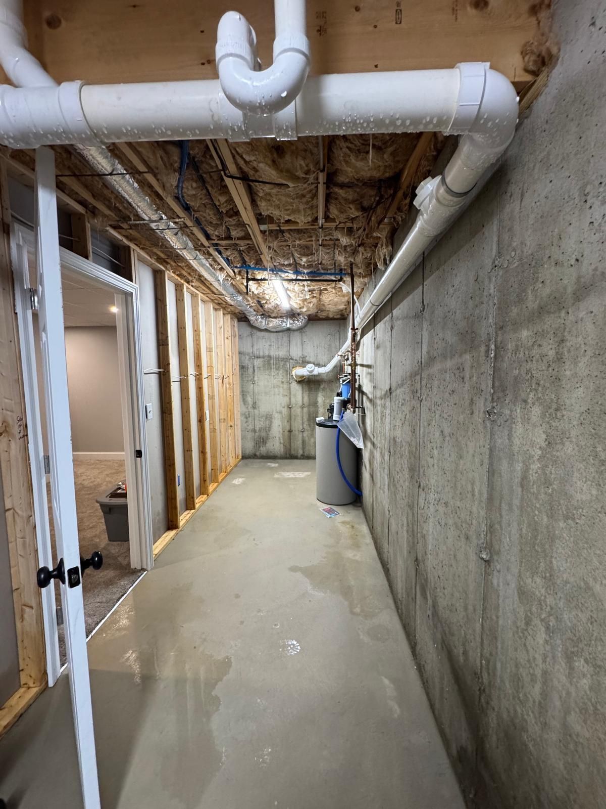 Narrow basement utility corridor with exposed ceiling pipes, concrete walls, and construction materials.