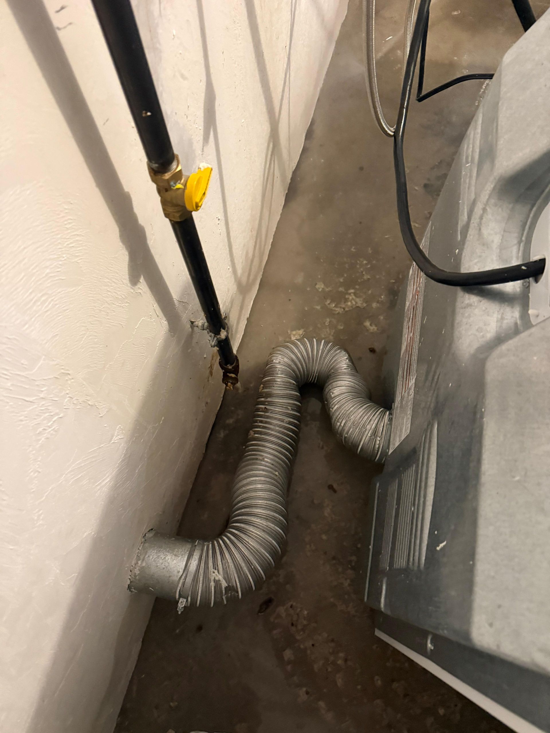 Flexible silver duct on floor beside wall pipes and a gray appliance in a narrow utility area
