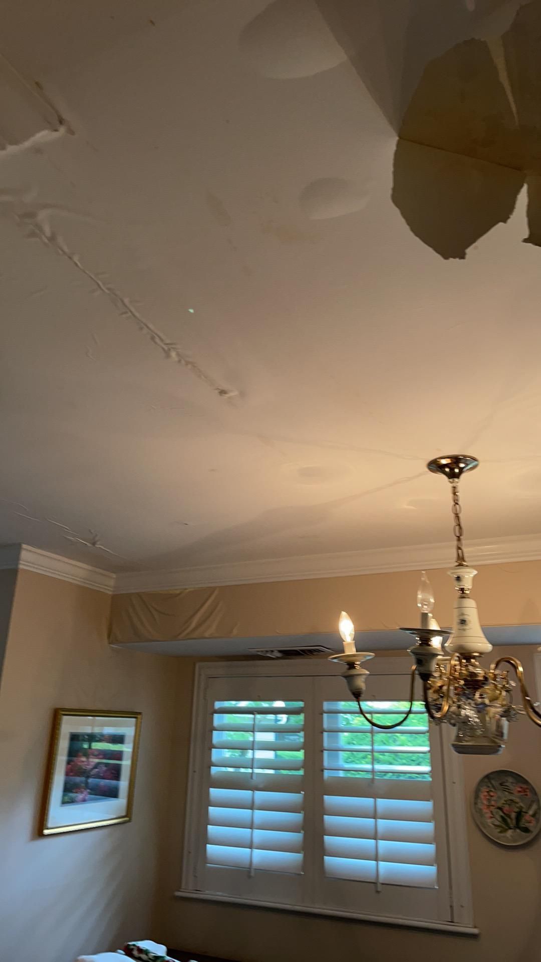 Ceiling with water damage and peeling paint above a window with white shutters and a hanging chandelier