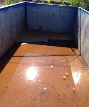 A rusty metal dumpster filled with murky brown water, some debris at the bottom, and bright sunlight reflecting on the surface.