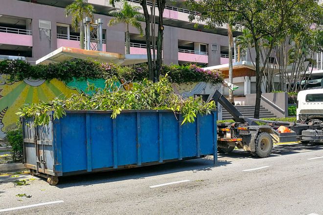 Blue dumpster filled with green branches being lifted by a truck on a street.