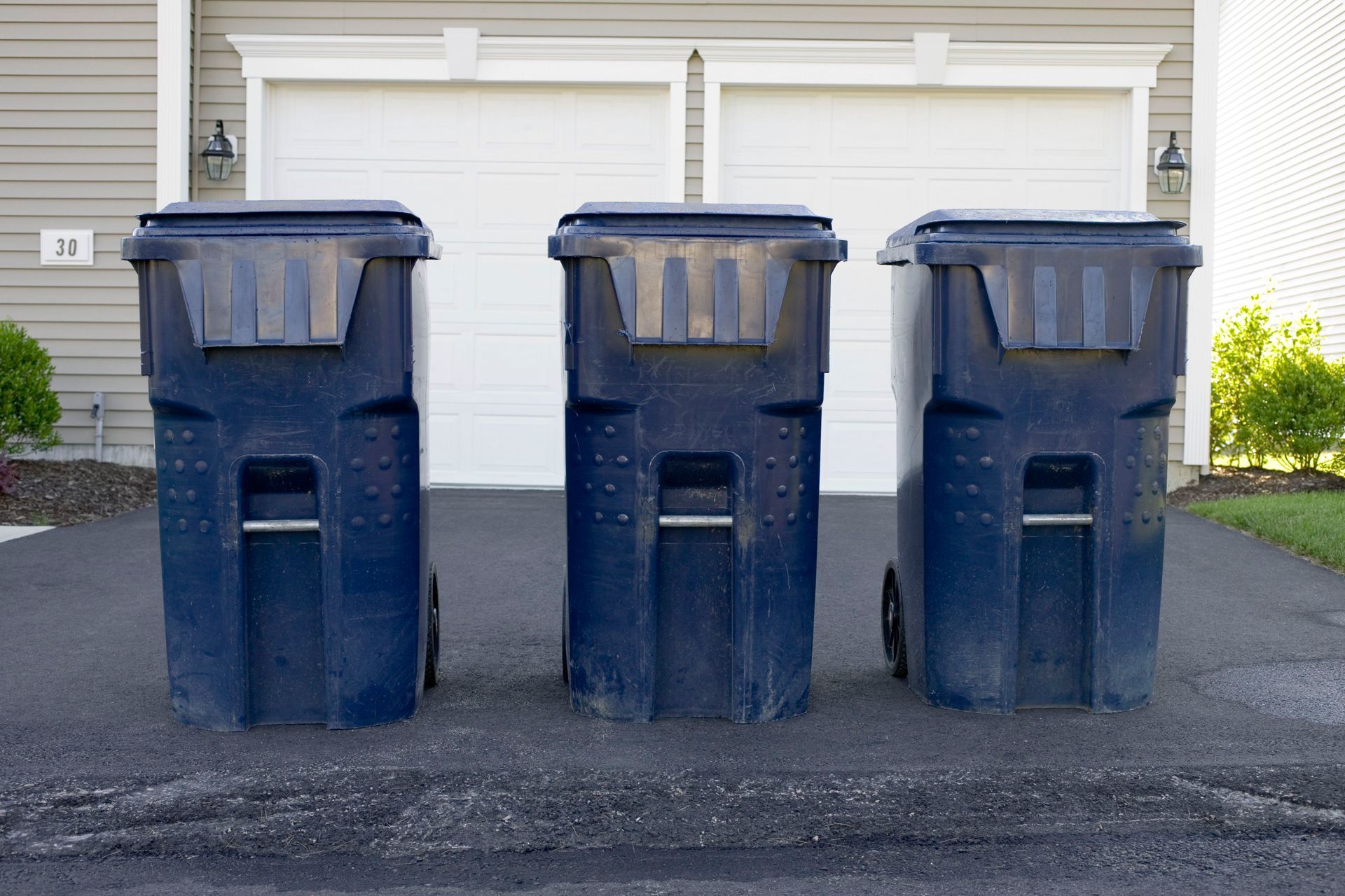 Three dark blue trash cans on a driveway in front of a garage.