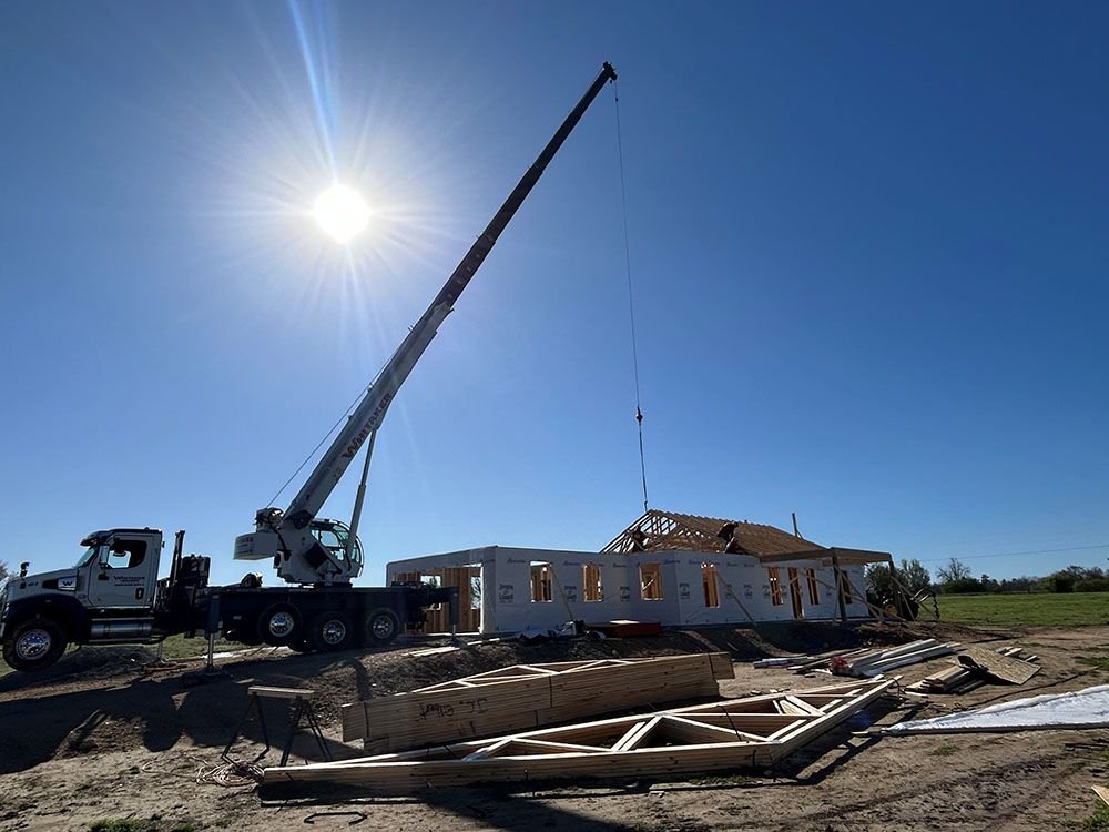 A Whitaker Towing, Hauling & Crane's crane lifts a roof truss onto the walls of a house under a clear blue sky.