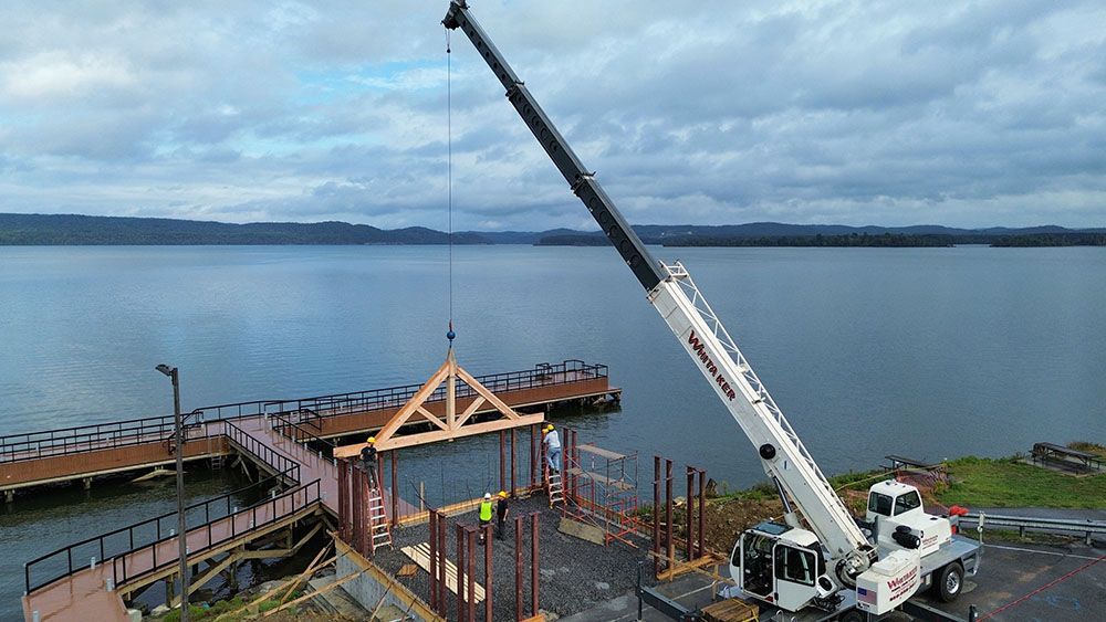 A Whitaker Towing, Hauling & Crane's lifts a wooden roof truss over a construction site on a pier extending into a lake under a cloudy sky.