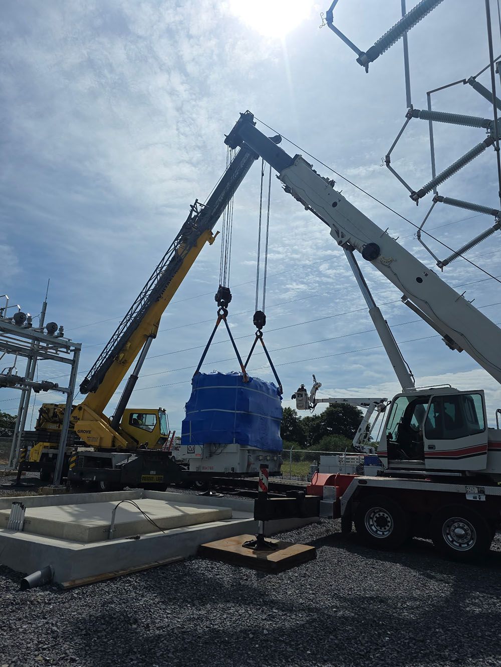 Two Whitaker Towing, Hauling & Crane's crane lifts a large, blue-wrapped piece of industrial equipment onto a concrete pad at a construction site.