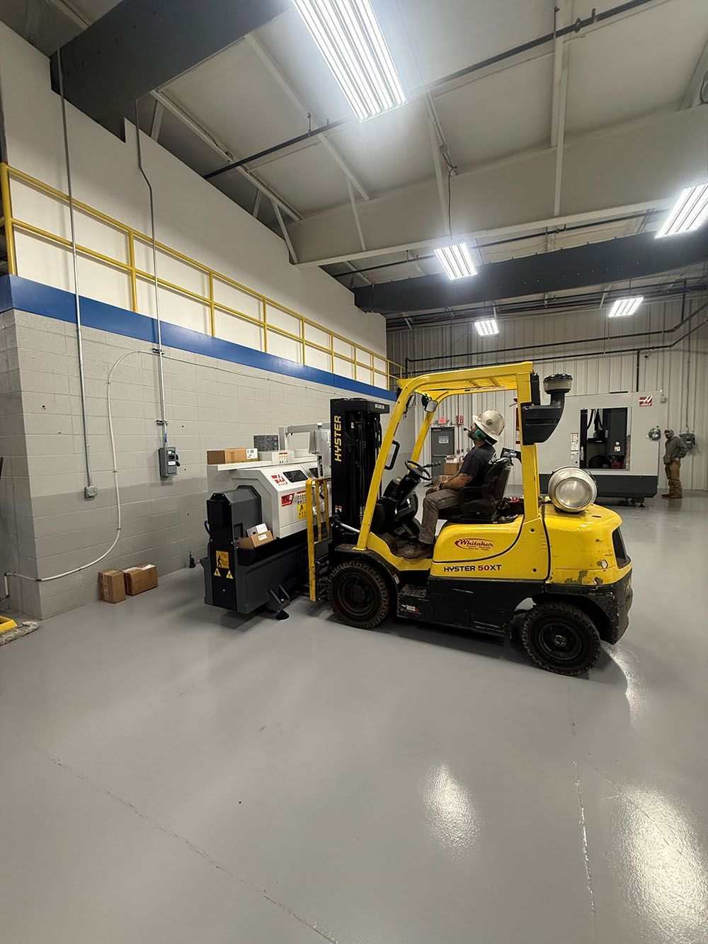 A person operates a Whitaker Towing, Hauling & Crane's forklift moving a piece of industrial machinery inside a warehouse with light gray flooring.