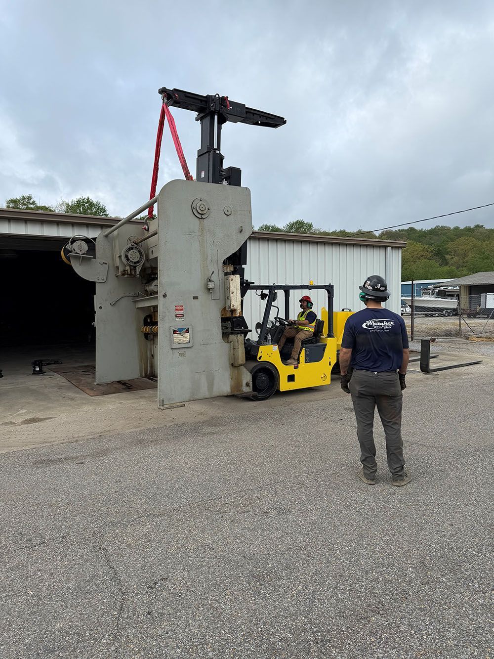 A worker watches a yellow Whitaker Towing, Hauling & Crane's forklift move a large industrial machine outside a storage building.