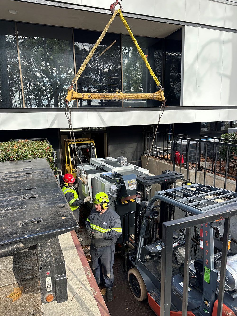 Two workers in high-visibility gear guide a heavy industrial machine being lifted by a Whitaker Towing, Hauling & Crane's crane outside a modern building.