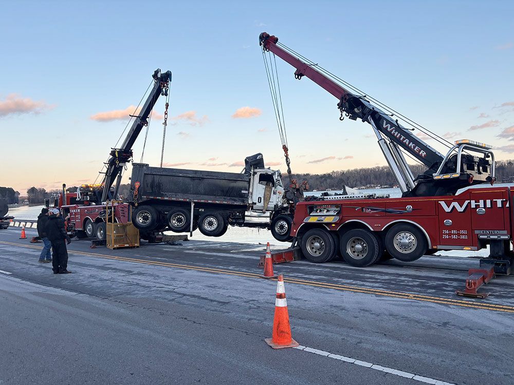 Two Whitaker Towing, Hauling & Crane tow trucks lift a black dump truck off the ground on an icy road at dusk.