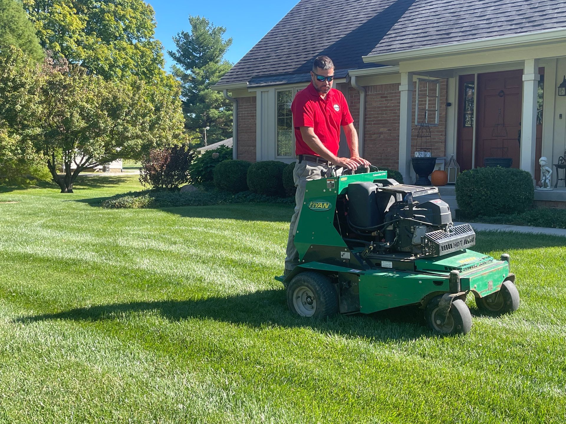 Man aerating a lawn with a green Bobcat machine in front of a house on a sunny day.