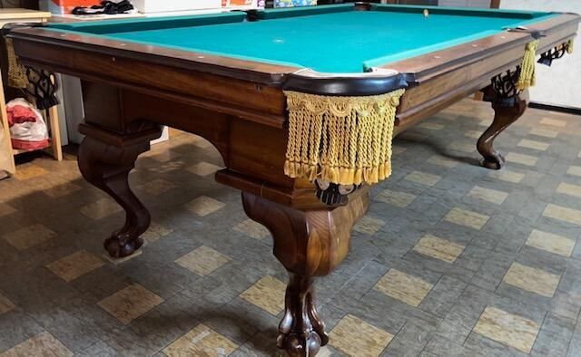 A wooden pool table with green felt on a colorful rug. Decorative fringe hangs at the pockets.