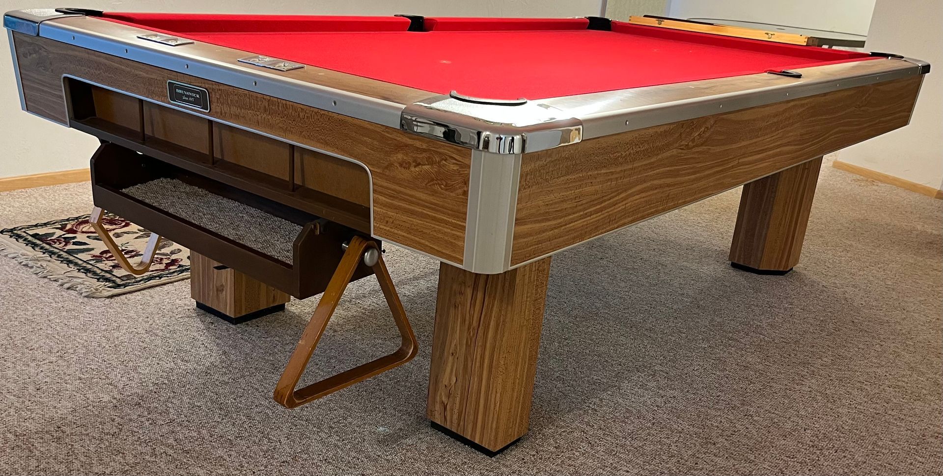 Red-felt pool table with wooden frame and legs in a carpeted room