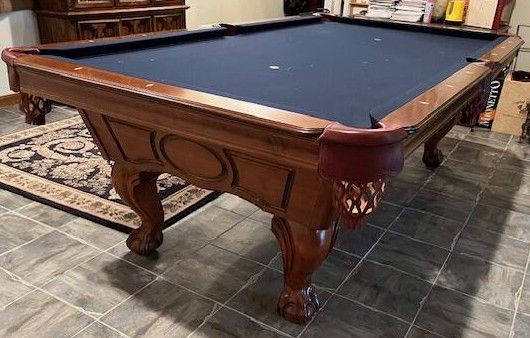 Wooden pool table with blue felt, ornate legs, and a patterned rug on a tiled floor.