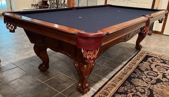 A brown, ornate pool table with dark blue felt, in a tiled room with a rug.