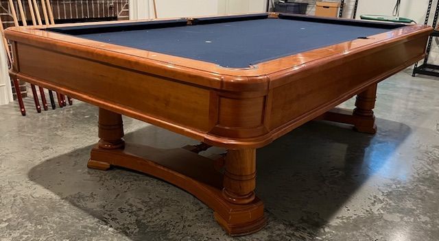 Wooden pool table with blue felt, in a room with concrete floor.