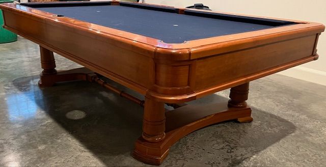 Wooden pool table with a black felt surface, set in a room with a concrete floor.
