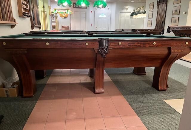 A large, dark wood vintage pool table with green felt, centered in an indoor room with tiled flooring and green lighting.
