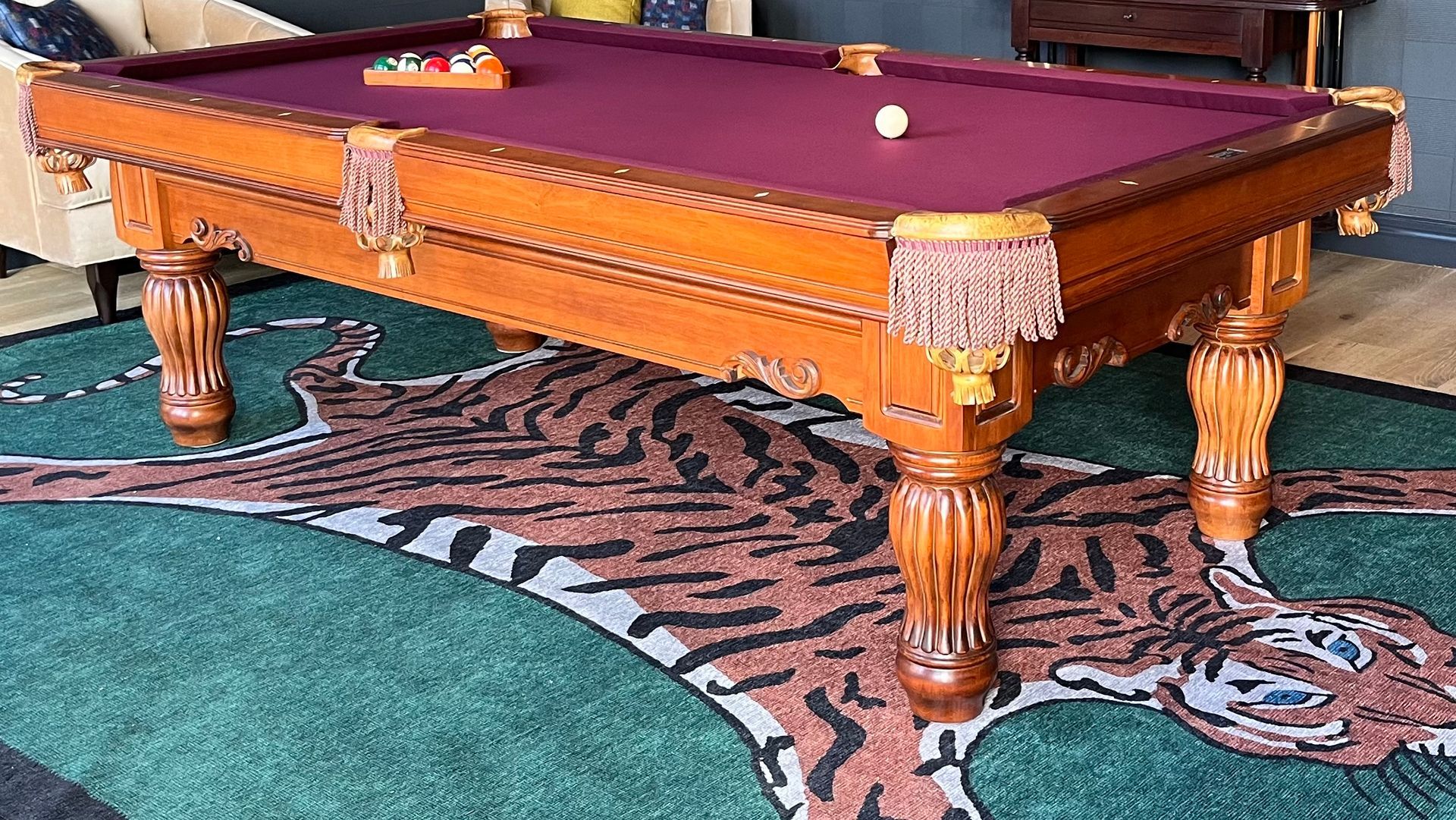 Pool table with burgundy felt and carved wooden legs, on a tiger-print rug.