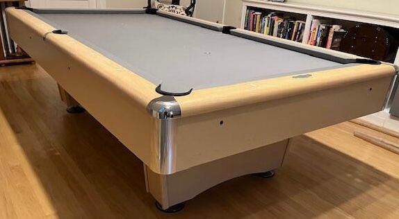 A tan and gray pool table on a hardwood floor, with books in the background.