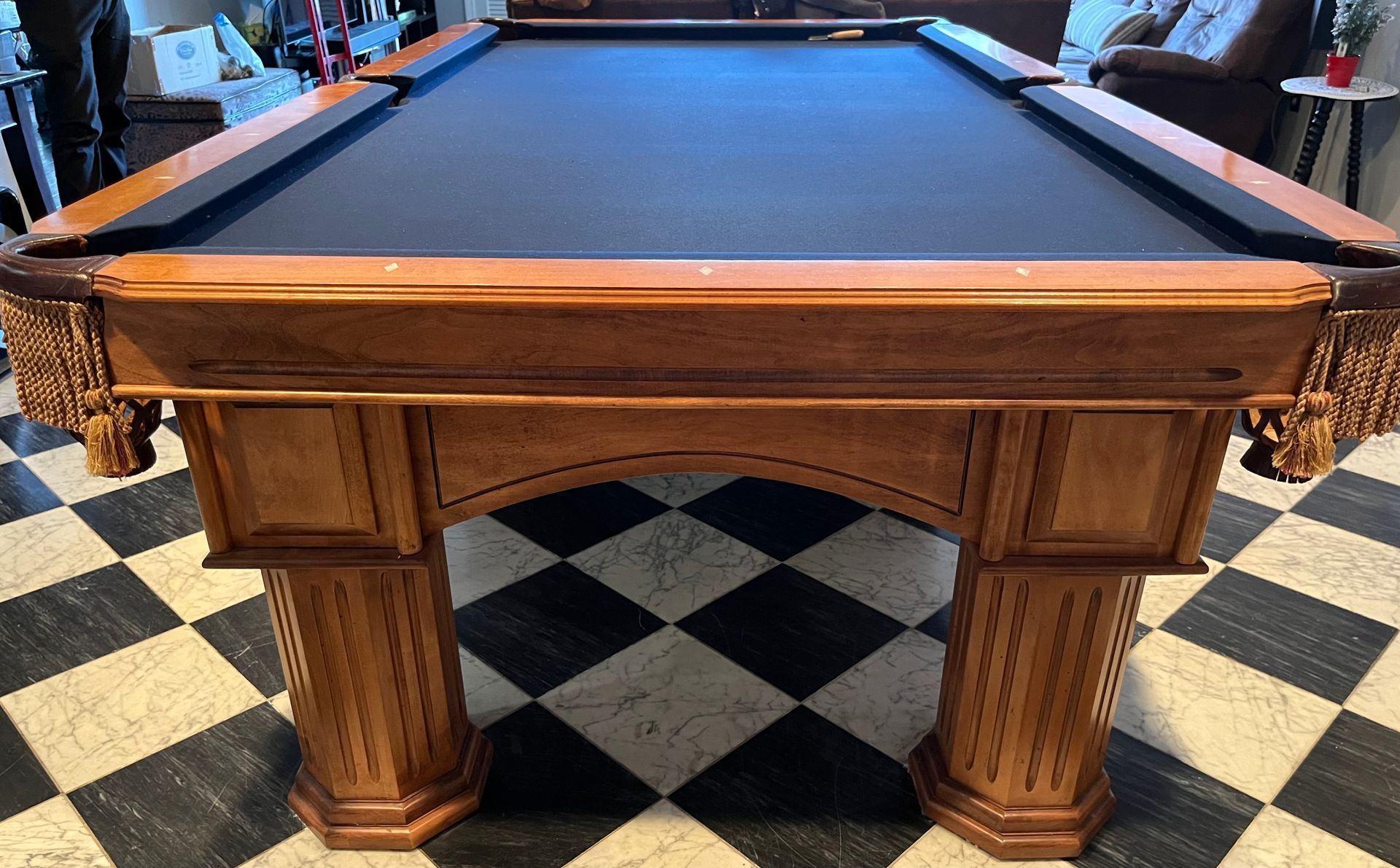 Wooden pool table with blue felt, tassel pockets, on a black and white tiled floor.