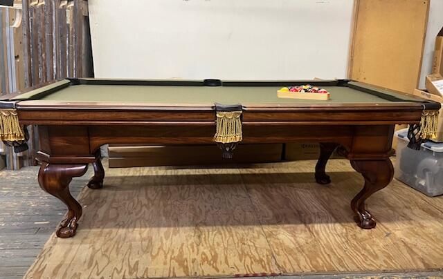 A wooden pool table with a green felt surface and gold-fringed pockets, standing on a plywood-covered floor.