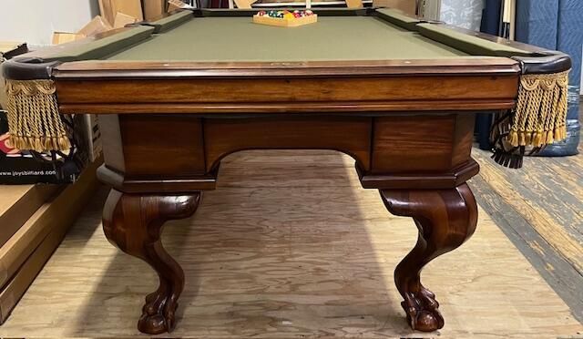 A wooden pool table with a green felt surface and decorative claw-and-ball feet featuring fringed corner pockets.