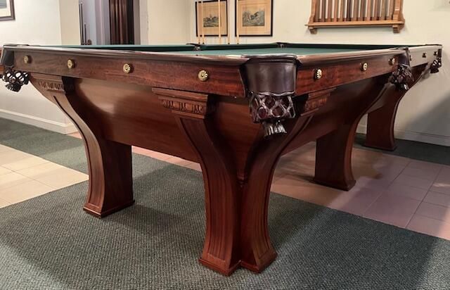 A dark wood antique-style pool table with carved legs and leather drop pockets stands on a green carpet.