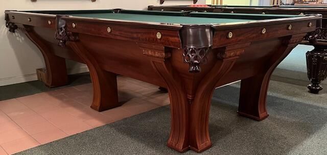 A vintage wooden pool table with green felt, leather-webbed corner pockets, and ornate carved legs in a showroom.