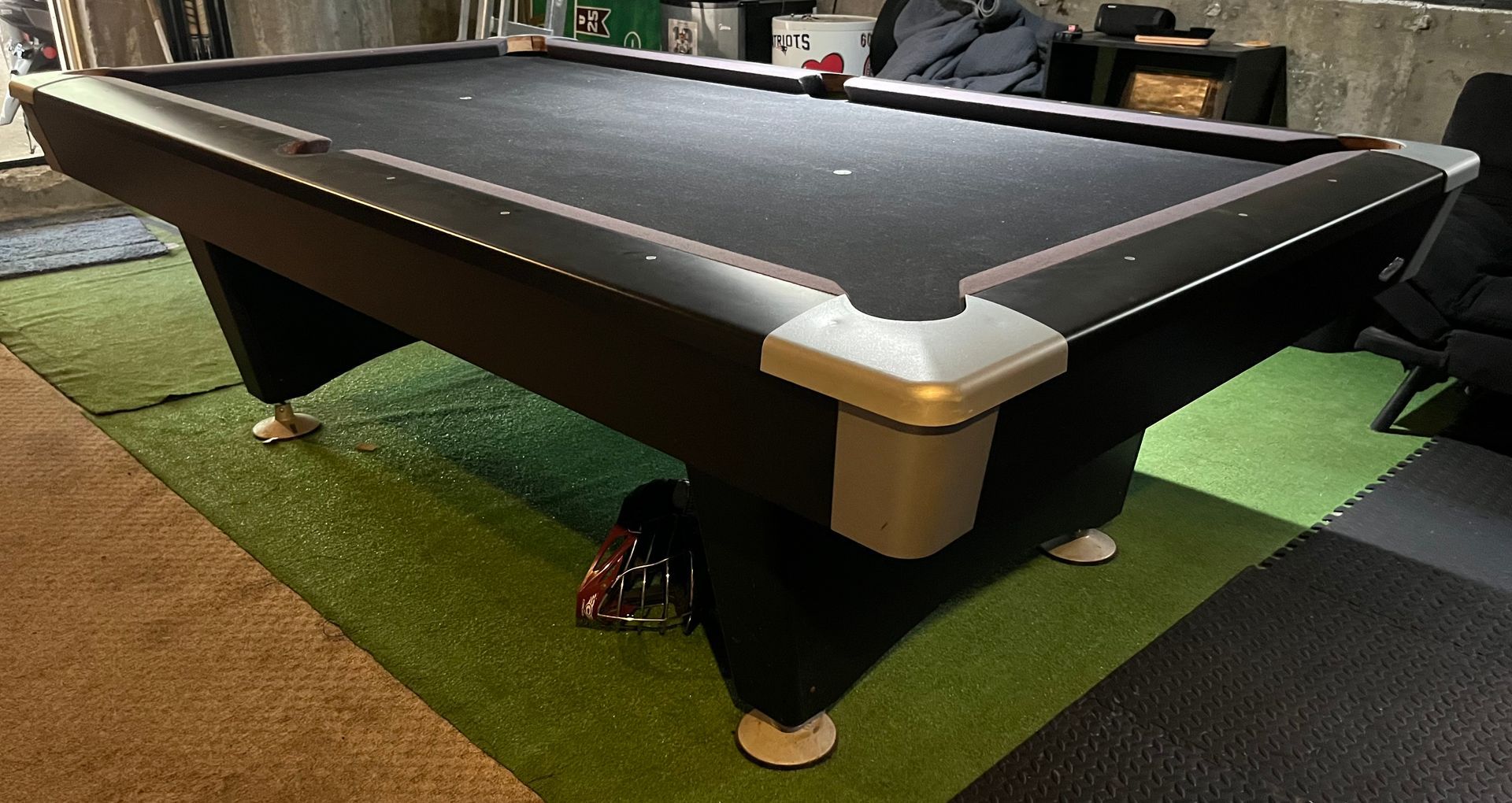 A pool table in a game room with black felt, tan rails, and green carpet underneath.