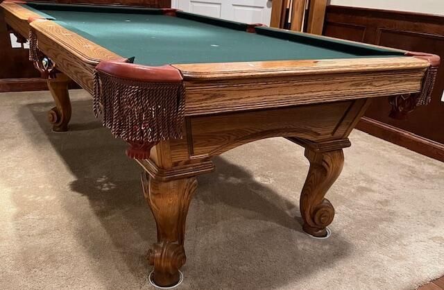 Wooden billiards table with green felt and red fringe pockets in a carpeted room