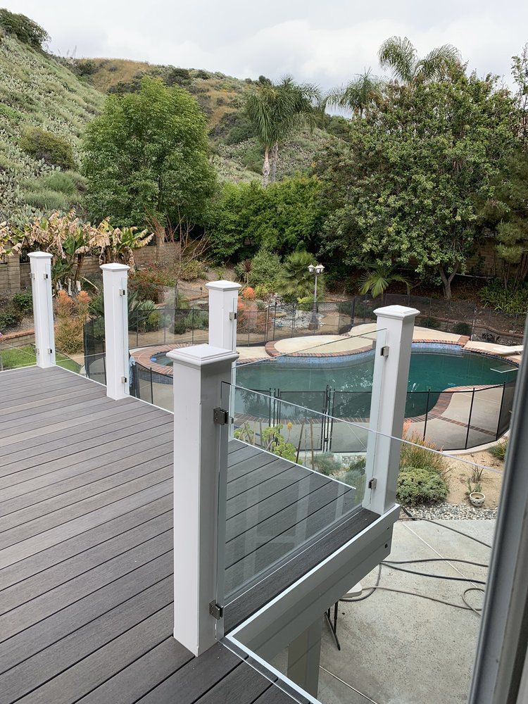 A deck with a glass railing and a pool in the background.