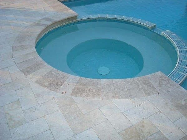 A circular hot tub with light blue water and stone coping sits beside a swimming pool on a light-colored stone patio.