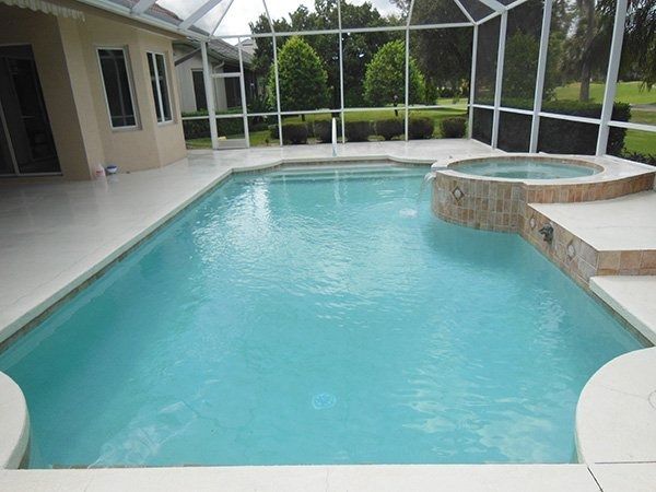 A rectangular swimming pool with an attached spillover spa inside a screened lanai enclosure next to a beige house.