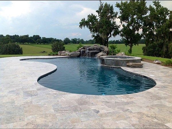 A curved swimming pool with a stone waterfall and hot tub, surrounded by a light-colored stone patio in a grassy yard.