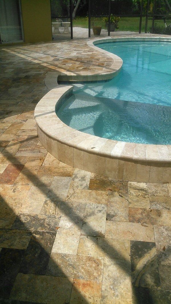 A curved concrete pool step area features light tan stone pavers surrounding a bright blue pool on a sunny day.