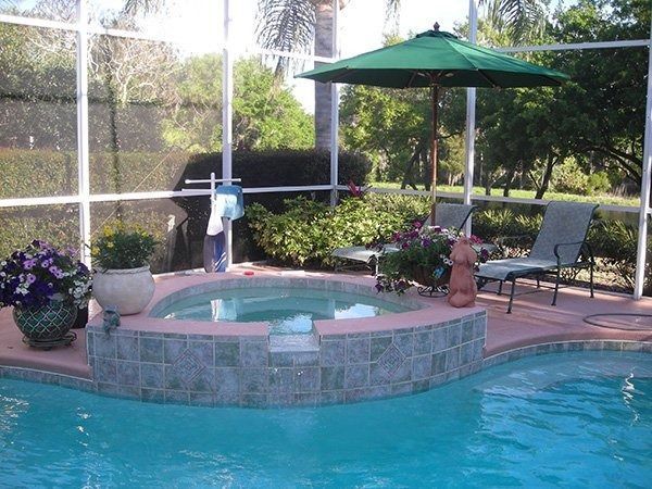 A hot tub spa built into a pool deck, with lounge chairs and a green umbrella under a screen enclosure.