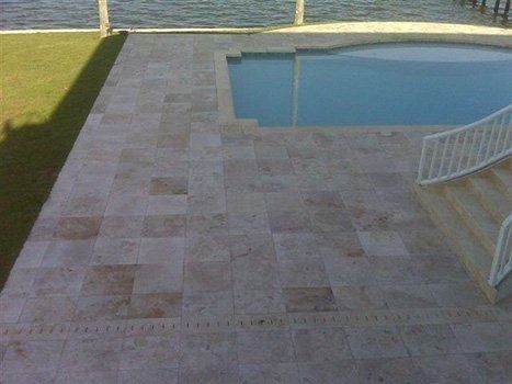 A patio paved with beige, rectangular stone tiles beside a swimming pool overlooking a body of water.
