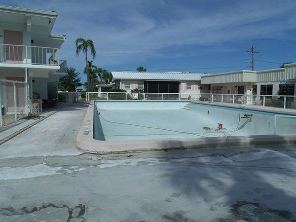 An empty swimming pool in front of a motel building under a bright blue sky.