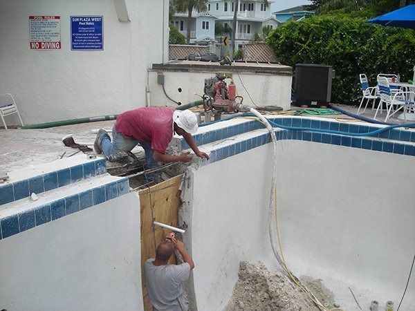 Two workers repair a tiled pool edge, with one kneeling on the deck and the other installing a board inside the drained pool.