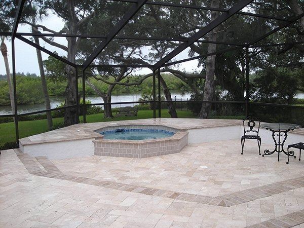 A screened-in patio featuring a raised circular hot tub, stone pavers, and a small bistro set overlooking a river.