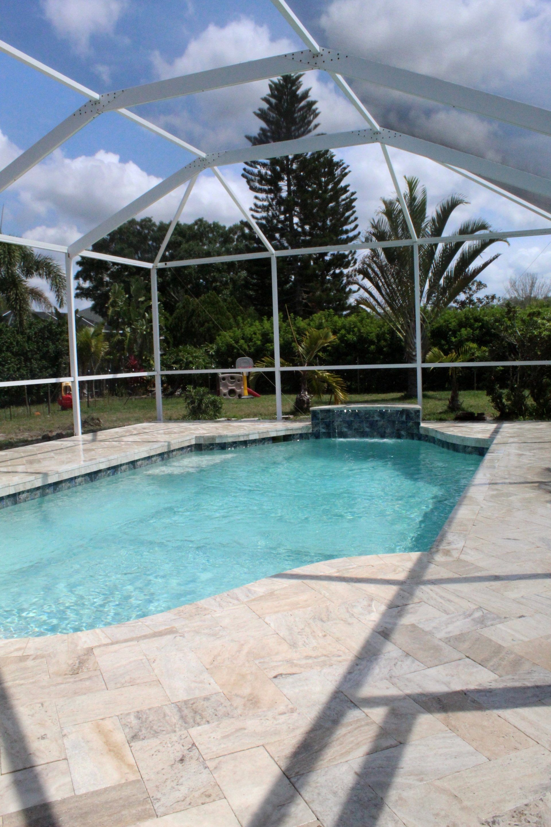 A backyard swimming pool with light-colored stone decking, enclosed by a white screen cage under a partly cloudy blue sky.