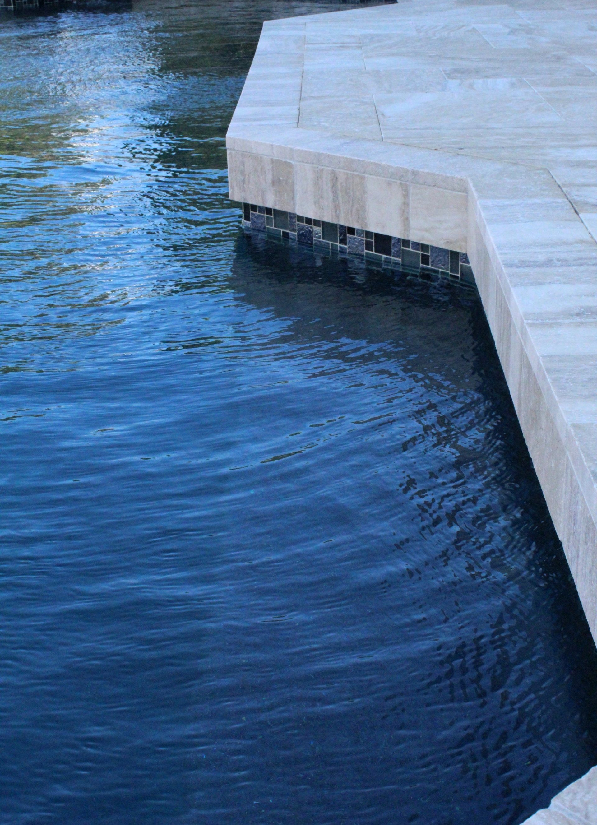 A stone-tiled pool deck with a cantilevered edge over deep blue, rippling water.