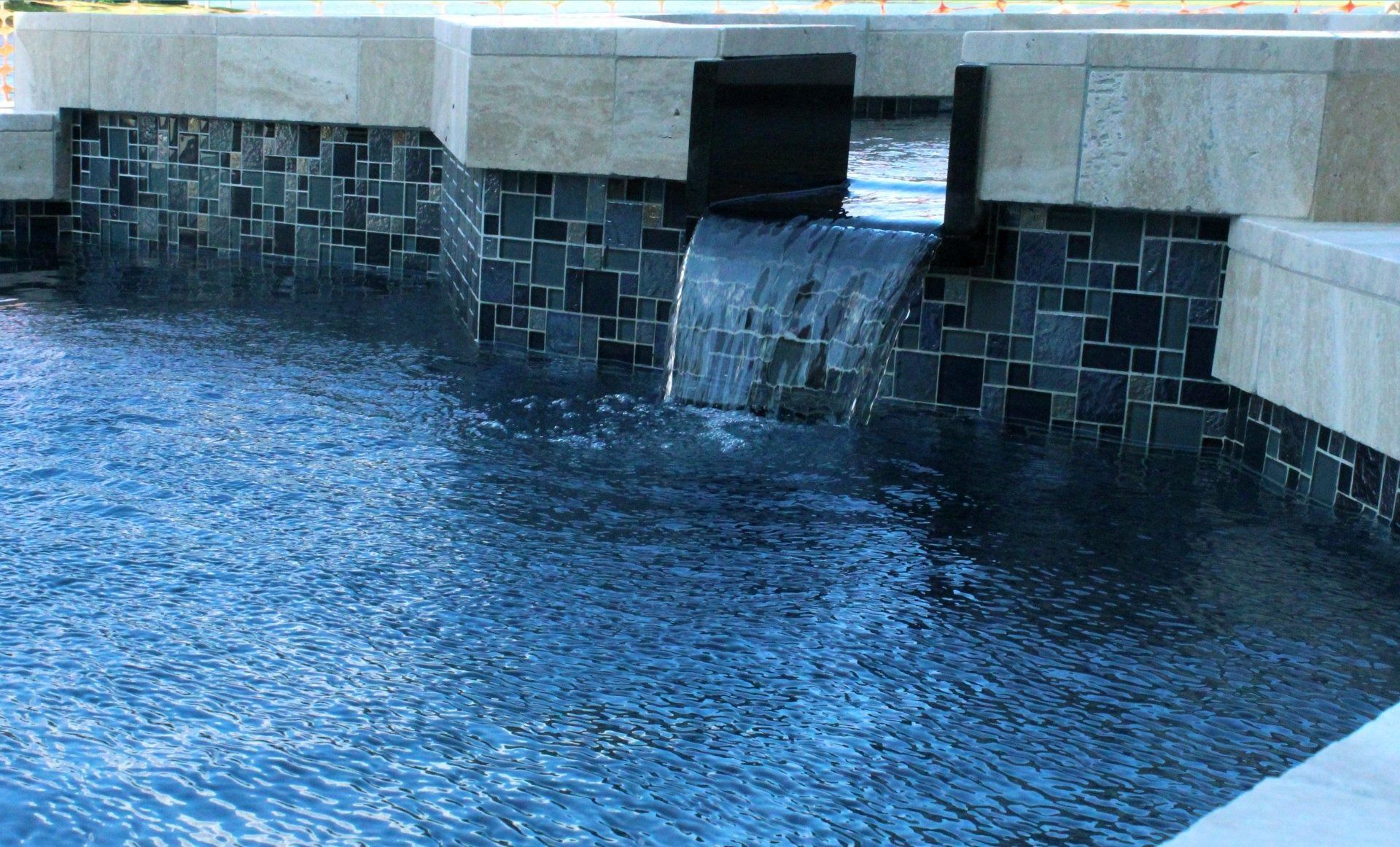 A waterfall feature pouring into a swimming pool with dark mosaic tiles and light-colored stone coping.